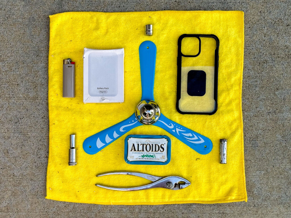 Blue boomerang with a plastic nut in the center, three sockets, pliers, empty Altoids tin, BIC lighter, magnetic battery pack, iPhone case on a yellow microfiber rag